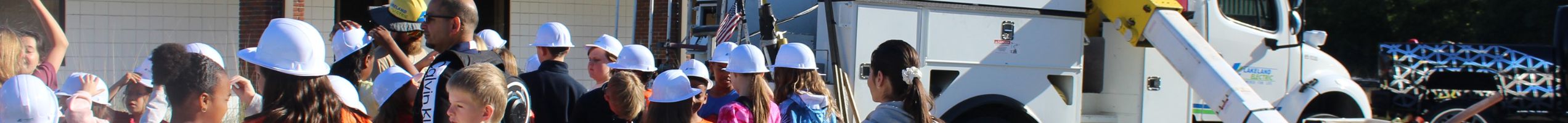 A group of Students, many wearing white hard hats, stand in front of a Lineman as he explains what he does beside a white bucket truck with a yellow crane arm.