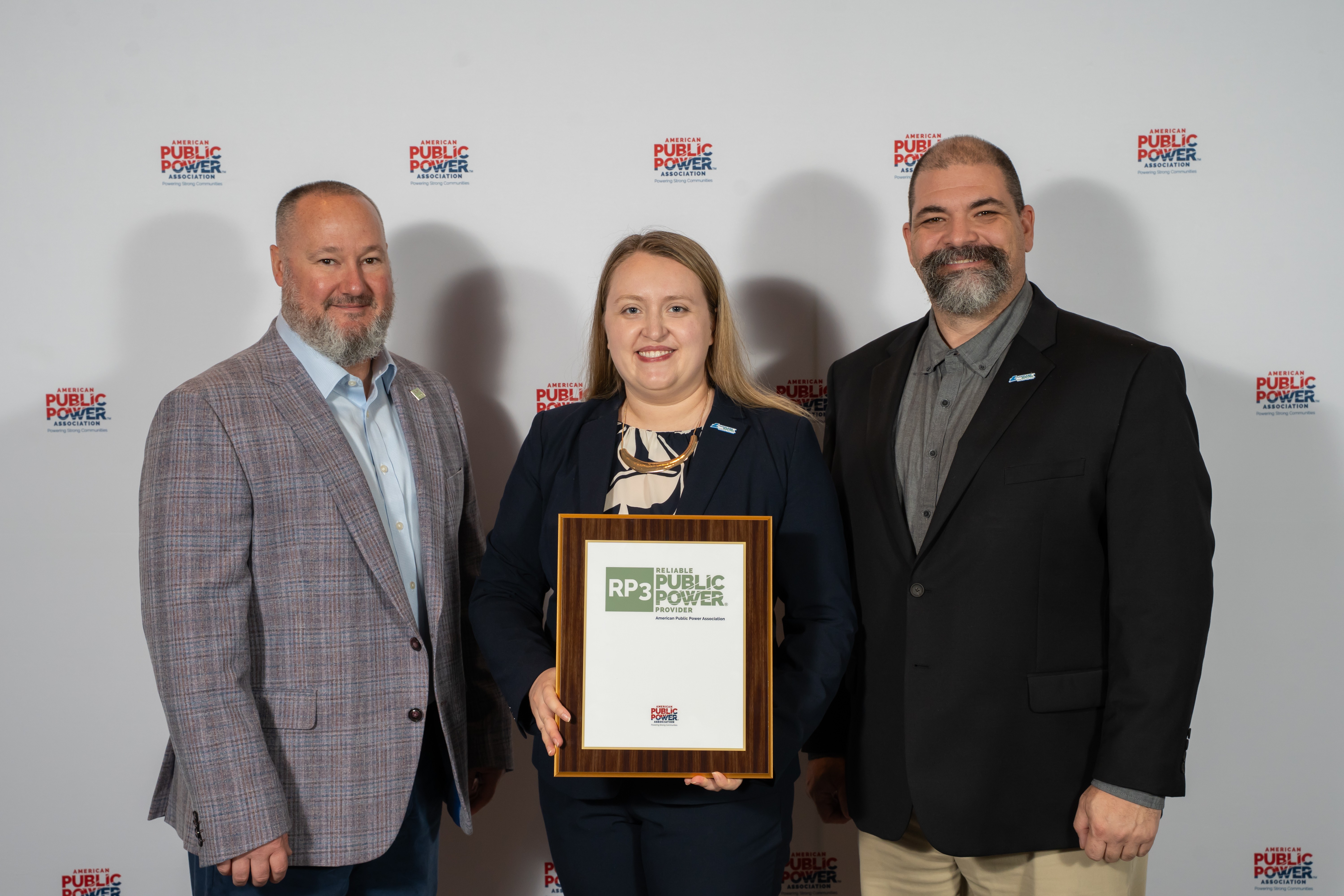 Three people stand in front of a backdrop with a logo. The person in the center holds a framed award. The person on the left wears a plaid jacket, and the person on the right wears a dark suit jacket.